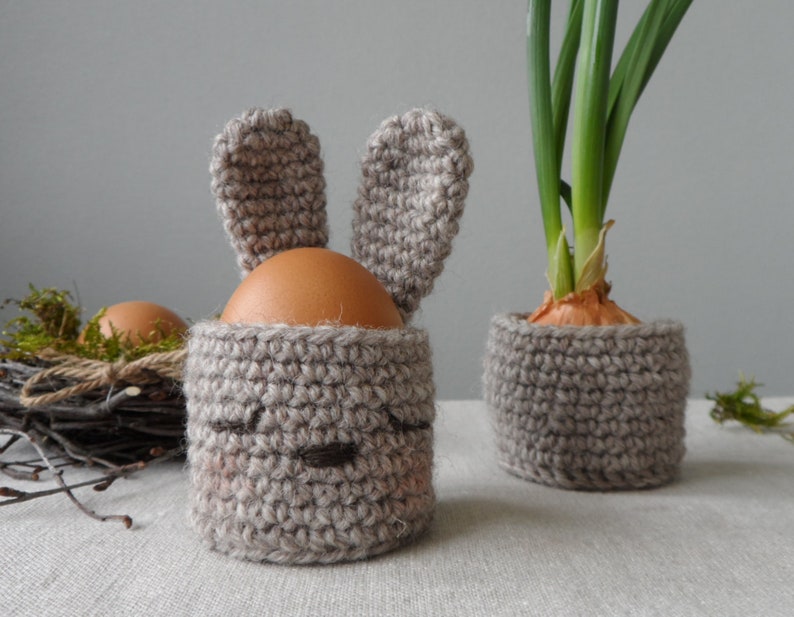 May include: Two crocheted bunny-shaped egg holders, one holding a brown egg and the other holding a potted onion plant with green shoots.