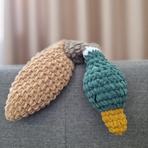 May include: A crocheted stuffed toy shaped like a duck. The duck is lying on its side with its head and neck resting on a gray couch. The duck is made of brown, green, and yellow yarn.