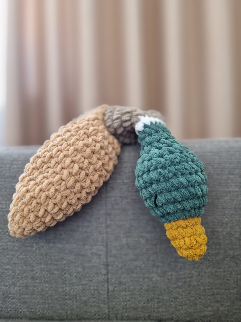 May include: A crocheted stuffed toy shaped like a duck. The duck is lying on its side with its head and neck resting on a gray couch. The duck is made of brown, green, and yellow yarn.