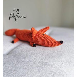 May include: A crocheted orange fox toy with black accents on the nose, paws, and eyes. The fox is lying down on a white surface. The words "PDF Pattern" are written in black script in the upper left corner.