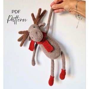 May include: A handmade crochet moose toy with brown antlers, a red scarf, and red feet. The moose is suspended by a long leg. The text "PDF Pattern" is in the upper left corner.