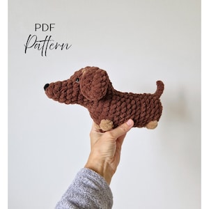 May include: A handmade brown crochet dachshund dog with tan paws and a black nose. The dog is held in a person's hand. The words "PDF Pattern" are written in cursive in the upper left corner.