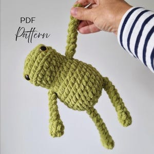 May include: A green crocheted frog toy with a textured yarn surface, dark round eyes, and long limbs. The text PDF Pattern is visible in the upper left corner. The frog is constructed with a thick, soft yarn and features a simple, rounded body shape.