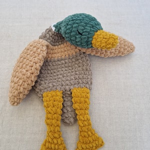 May include: A crocheted plush toy duck with a green head, brown body, and yellow legs. The duck is lying on its side with its head resting on its wing.