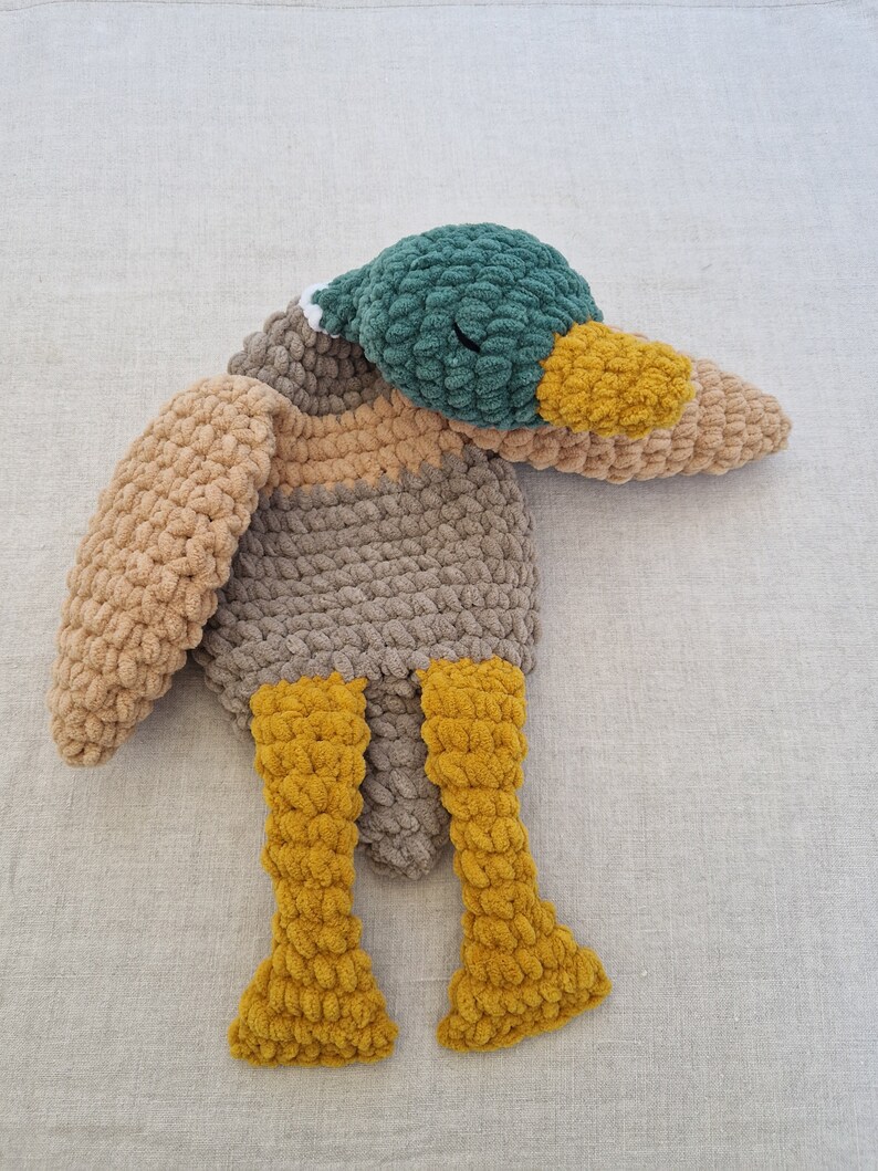 May include: A crocheted plush toy duck with a green head, brown body, and yellow legs. The duck is lying on its side with its head resting on its wing.
