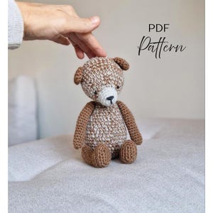 Op de afbeelding: Een gehaakte teddybeer in bruin- en wittinten, met een zwarte neus en kleine oren. De beer zit op een lichtgrijze ondergrond. De tekst "PDF Pattern" is zichtbaar.