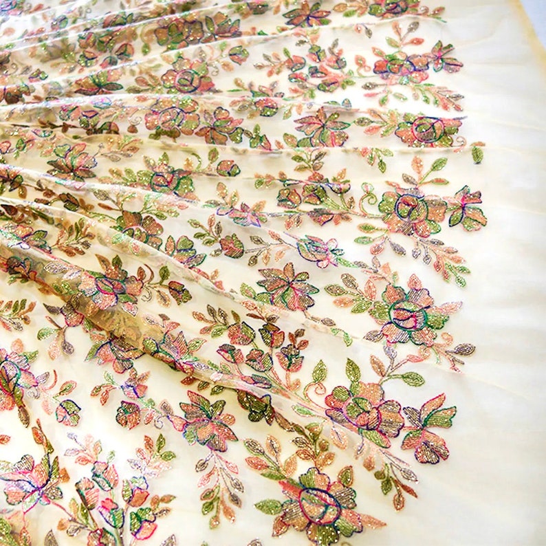 May include: White sheer fabric with a floral pattern embroidered with iridescent sequins. The flowers are in shades of pink, green, and brown.