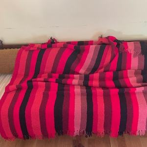 Könnte beinhalten: Eine lebendige, gestreifte Decke in Rot-, Rosa- und Schwarztönen mit einem Fransenrand. Die Decke ist über einer Holzbank mit einem Polsterkissen drapiert. Die Farbpalette erzeugt eine warme und einladende Ästhetik.