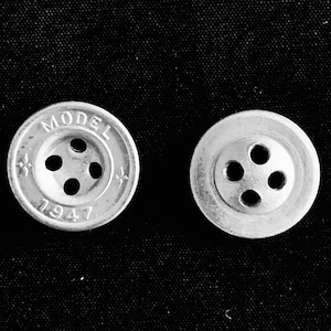 May include: Two silver-colored buttons with four holes each, set against a black background. One button has the word "MODEL" and the number "1947" inscribed on it. The buttons are round with a slightly raised edge.
