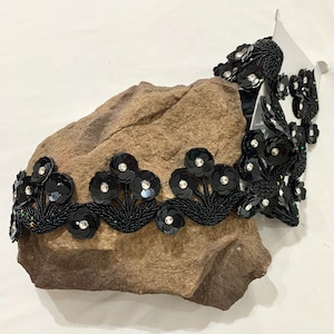 May include: Black beaded trim with sequined flower embellishments. The trim is on a brown rock surface.