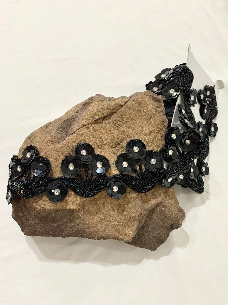 May include: Black beaded trim with sequined flower embellishments. The trim is on a brown rock surface.
