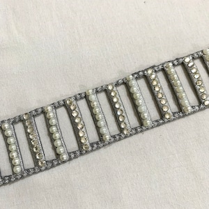 Pode incluir: Uma guarnição prateada com fileiras de strass transparentes e pérolas brancas. A guarnição é projetada para ser costurada em tecido.