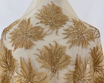 May include: A sheer, beige fabric featuring a repeating pattern of gold floral embroidery. The floral design is embellished with sequins, creating a shimmering effect. The fabric is draped over a mannequin, highlighting its texture and drape.