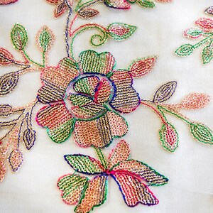 May include: Close-up of a white fabric with embroidered floral designs. The flowers are made of sequins in shades of pink, green, gold, and blue.