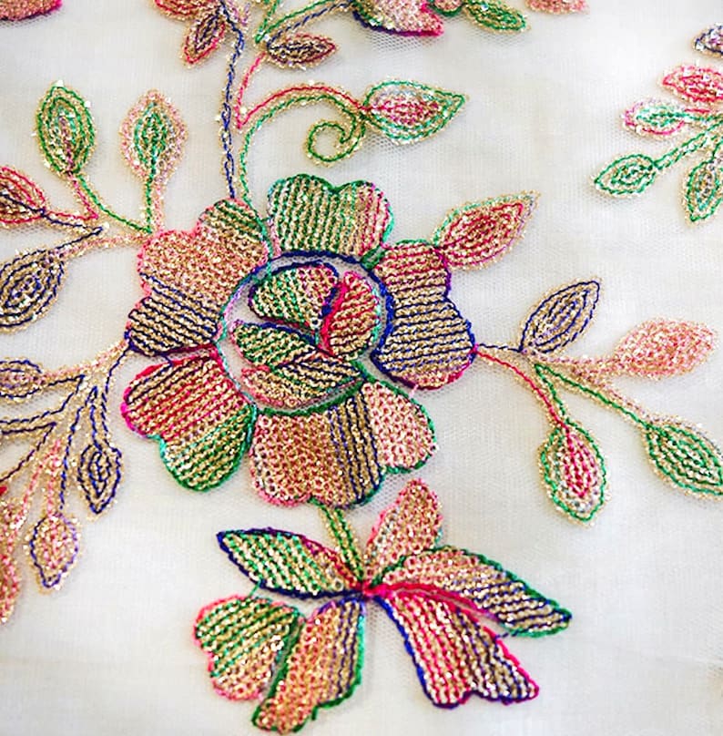 May include: Close-up of a white fabric with embroidered floral designs. The flowers are made of sequins in shades of pink, green, gold, and blue.