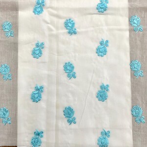 May include: White fabric with a light blue floral embroidery pattern. The fabric is folded in half and the embroidery is visible on both sides.