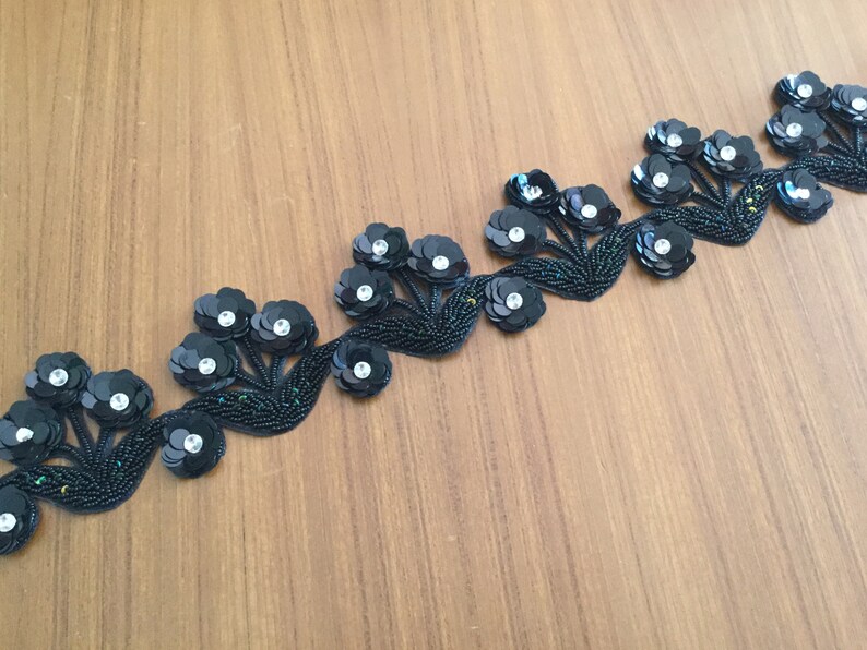 May include: Black beaded trim with a repeating pattern of floral designs. Each flower is made of black sequins with a clear rhinestone center.