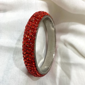 Può includere: Un braccialetto rosso con strass con una fascia in metallo color argento. Il braccialetto è ricoperto di piccoli strass rossi scintillanti. Il gioiello è posto su uno sfondo bianco.