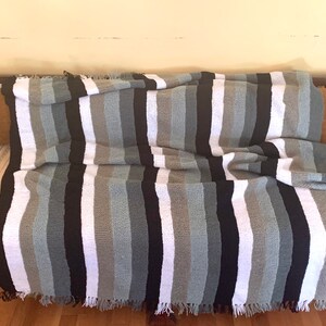 May include: A striped throw blanket in shades of white, gray, and black with fringe on the edges. The blanket is draped over a wooden couch.