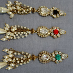 May include: Three ornate, gold-toned jewelry pieces with pearl and crystal accents. Each piece features a teardrop-shaped pearl cluster, a chain of small pearls, and a decorative centerpiece with a large, clear crystal and smaller surrounding crystals. One has a black accent, one a red rose, and one a teal rose.