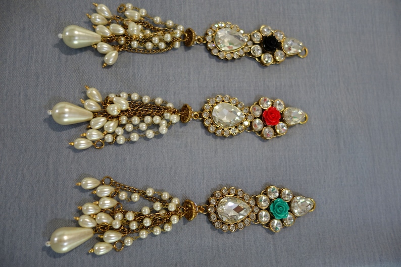 May include: Three ornate, gold-toned jewelry pieces with pearl and crystal accents. Each piece features a teardrop-shaped pearl cluster, a chain of small pearls, and a decorative centerpiece with a large, clear crystal and smaller surrounding crystals. One has a black accent, one a red rose, and one a teal rose.