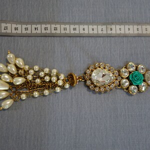 May include: An ornate gold-toned brooch with a teardrop-shaped clear crystal and a cluster of faux pearls. The brooch features a turquoise rose and clear crystals. The brooch measures approximately 7 inches in length.