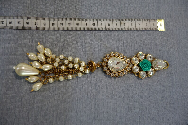 May include: An ornate gold-toned brooch with a teardrop-shaped clear crystal and a cluster of faux pearls. The brooch features a turquoise rose and clear crystals. The brooch measures approximately 7 inches in length.