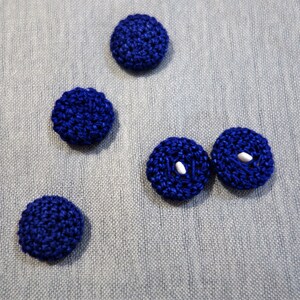 Puede incluir: Cinco botones de crochet azul con centros blancos. Los botones están dispuestos en semicírculo sobre un fondo de tela azul.