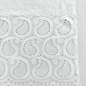 May include: White cotton fabric with a delicate embroidered border featuring a paisley pattern. The border is made of small, intricate stitches and has a scalloped edge.