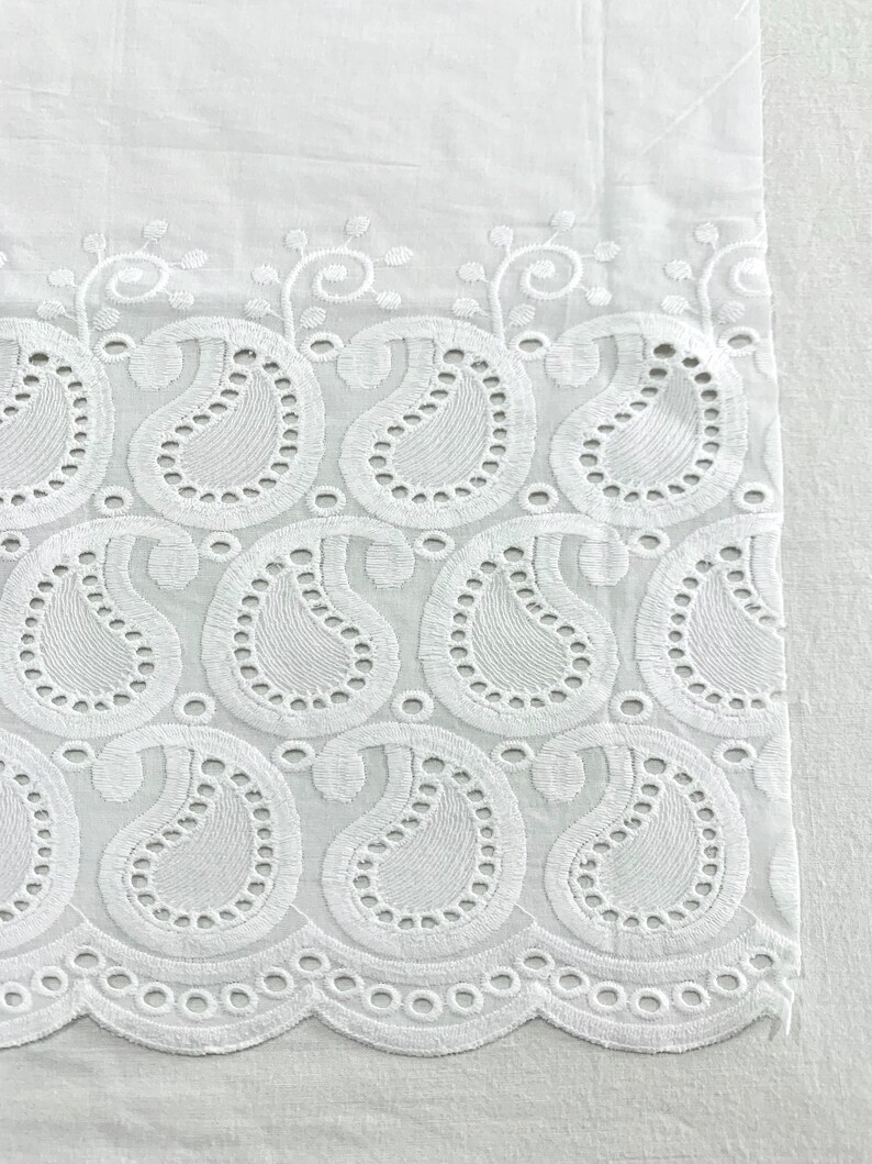 May include: White cotton fabric with a delicate embroidered border featuring a paisley pattern. The border is made of small, intricate stitches and has a scalloped edge.