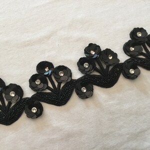 May include: Black beaded trim with sequined flower embellishments. The trim features a repeating pattern of five-petal flowers with a central rhinestone.