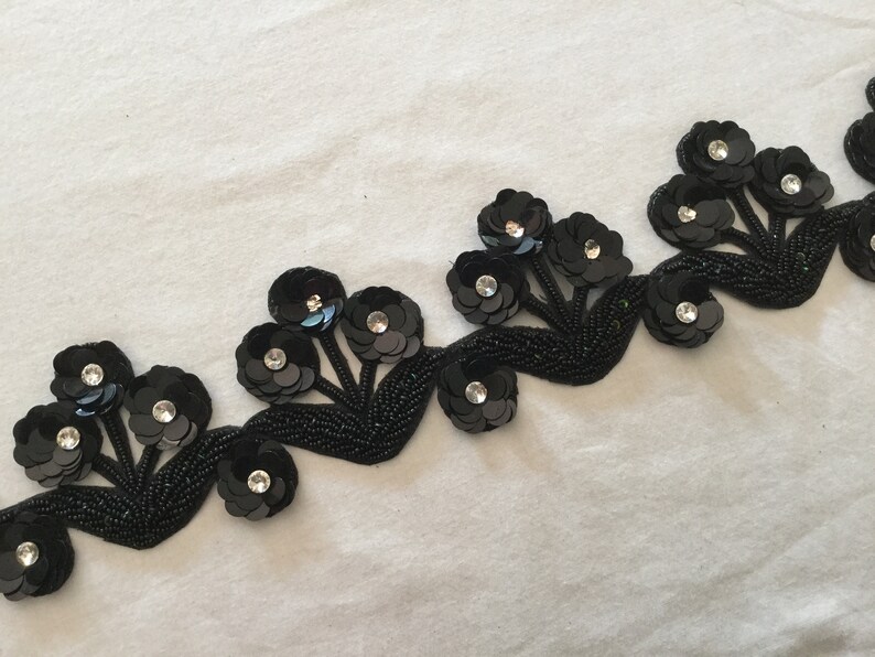 May include: Black beaded trim with sequined flower embellishments. The trim features a repeating pattern of five-petal flowers with a central rhinestone.