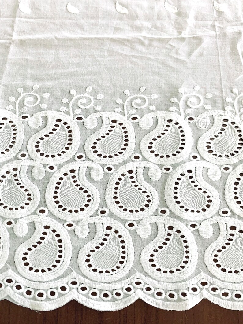 May include: White cotton fabric with a delicate embroidered border featuring a paisley pattern. The border has a scalloped edge with small circles.
