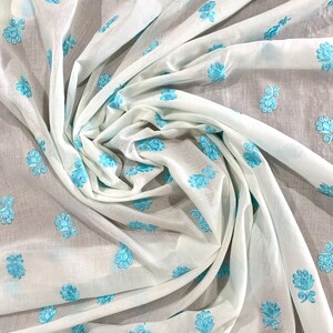 May include: White fabric with a light blue floral embroidery pattern. The fabric is folded and draped, showcasing the intricate design.