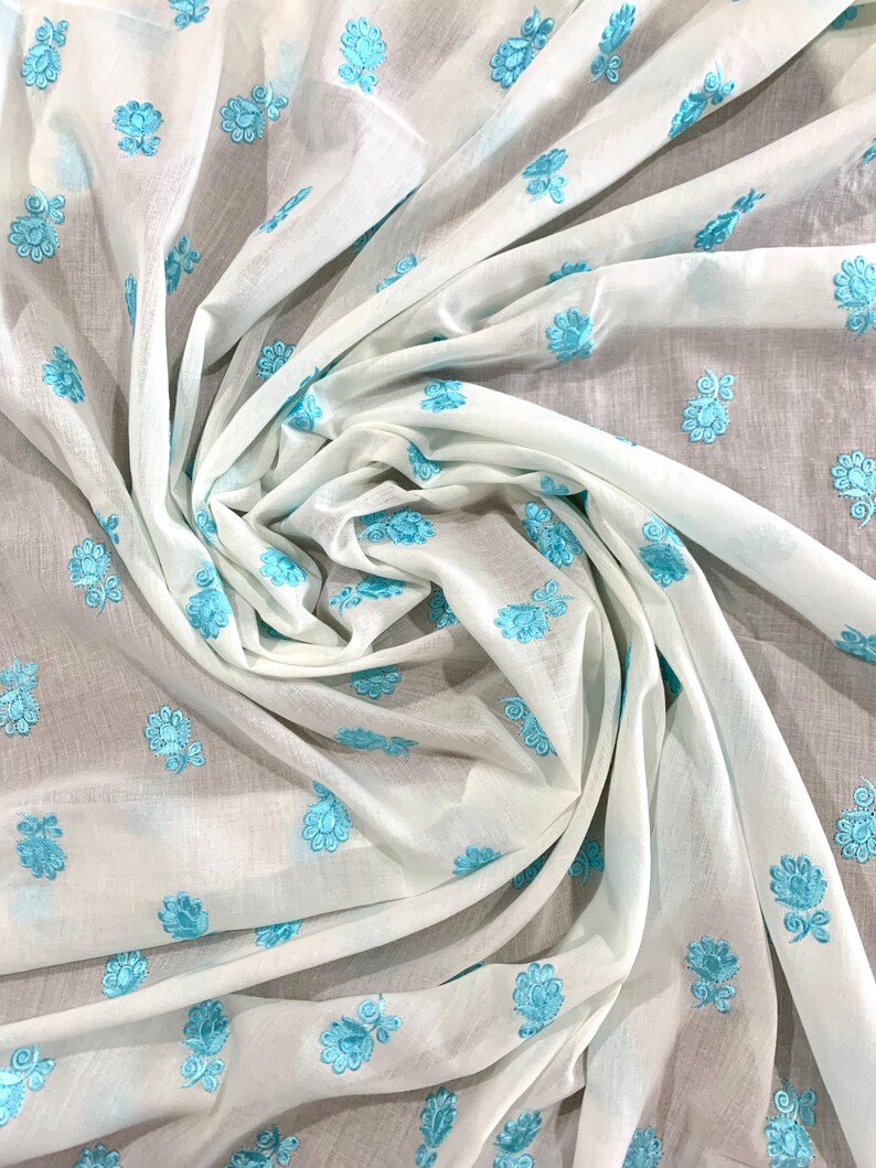 May include: White fabric with a light blue floral embroidery pattern. The fabric is folded and draped, showcasing the intricate design.