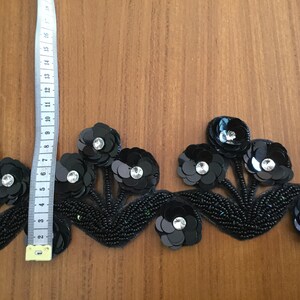 May include: Black beaded trim with black and silver sequins in a floral design. The trim is approximately 3 inches wide.