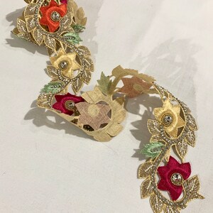 May include: Gold trim lace with floral embroidery and beaded accents. The lace features red, orange, and green flowers with clear rhinestones in the center.