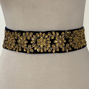 May include: A black velvet belt with gold floral embroidery. The belt is adorned with small, round, gold-colored embellishments.