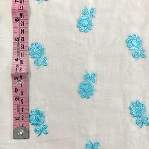 May include: White fabric with a repeating pattern of light blue embroidered flowers. The fabric is measured with a ruler in inches.
