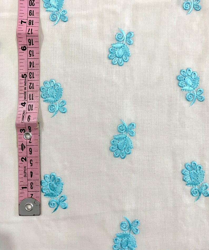 May include: White fabric with a repeating pattern of light blue embroidered flowers. The fabric is measured with a ruler in inches.