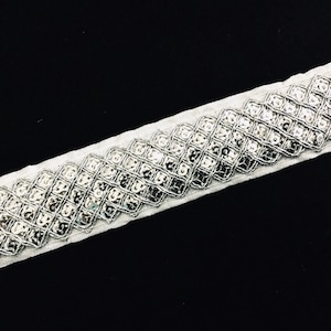 May include: Silver sequined trim with a white fabric backing. The sequins are arranged in a diagonal pattern.