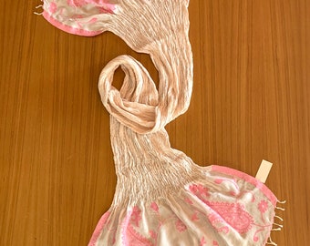 May include: A white and pink paisley patterned scarf with a fringed edge. The scarf is folded and draped on a wooden surface.