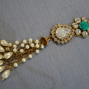 May include: An ornate, gold-toned jewelry piece with a teardrop-shaped clear gem and a cluster of smaller clear gems. A small turquoise rose is set among the gems. A tassel of faux pearls hangs from the piece.