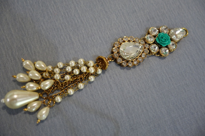 May include: An ornate, gold-toned jewelry piece with a teardrop-shaped clear gem and a cluster of smaller clear gems. A small turquoise rose is set among the gems. A tassel of faux pearls hangs from the piece.