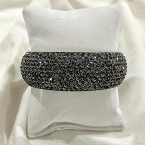 Puede incluir: Una pulsera rígida y ancha, completamente cubierta de pequeños cristales facetados de color gris oscuro. La superficie de la pulsera está densamente poblada con estas piedras reflectantes, creando una apariencia uniforme, texturizada y brillante en toda la banda exterior.