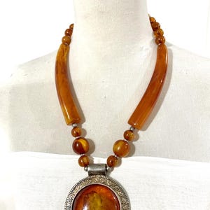 May include: A necklace featuring a large, round, amber-colored cabochon pendant set in a decorative silver-toned metal frame. The chain consists of smooth, translucent amber-colored beads and two elongated, curved amber-colored tube-shaped segments.