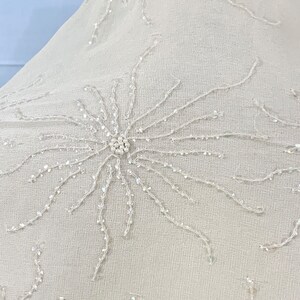 Pode incluir: Close-up de um tecido branco com um padrão delicado de flores de contas. As contas são pequenas e iridescentes, criando um brilho sutil.