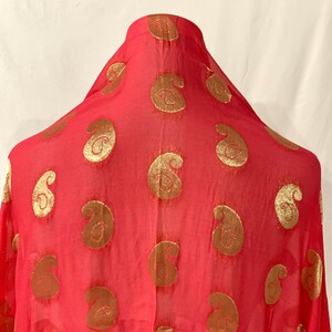 Pode incluir: Um lenço vermelho transparente com um padrão paisley dourado. O lenço está drapeado sobre a cabeça de um manequim.