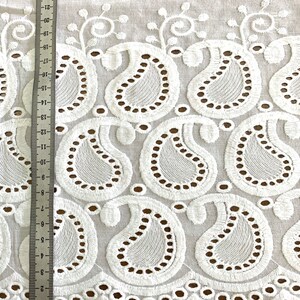 May include: White cotton fabric with a scalloped edge and an embroidered paisley pattern. The fabric is shown with a ruler measuring 22 inches.