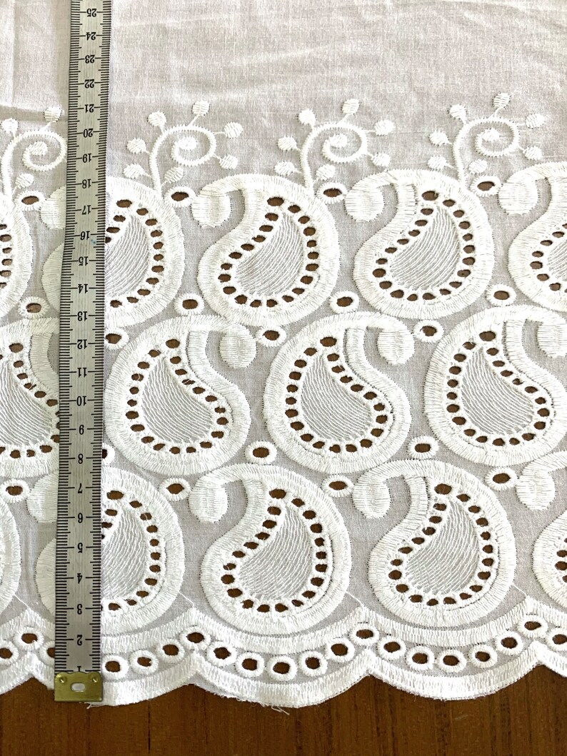 May include: White cotton fabric with a scalloped edge and an embroidered paisley pattern. The fabric is shown with a ruler measuring 22 inches.
